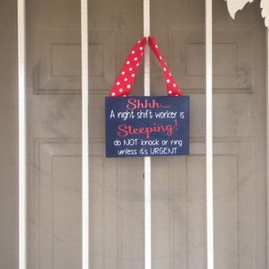 May include: A navy blue sign with white and red text that reads "Shhh.... A night shift worker is Sleeping! do NOT knock or ring unless it's URGENT." The sign is suspended by a red ribbon with white polka dots.