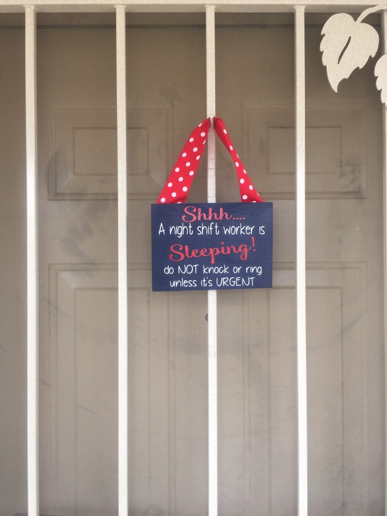 May include: A navy blue sign with white and red text that reads "Shhh.... A night shift worker is Sleeping! do NOT knock or ring unless it's URGENT." The sign is suspended by a red ribbon with white polka dots.