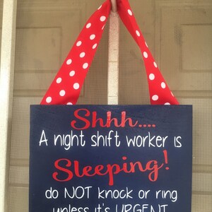 May include: A navy blue rectangular sign with white and red text, suspended by a red ribbon with white polka dots. The sign reads "Shhh... A night shift worker is Sleeping! do NOT knock or ring unless it's URGENT."