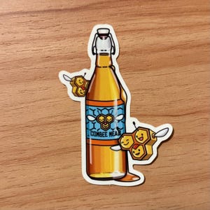 May include: A sticker featuring a bottle of Combee Mead with a white and silver stopper. The bottle is filled with a golden liquid and has a blue label with a honeycomb design. Two cartoon honeycombs with smiling faces are on either side.