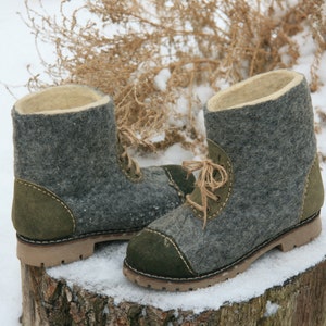 May include: A pair of gray felt boots with green suede accents and brown laces. The boots are sitting on a tree stump covered in snow.
