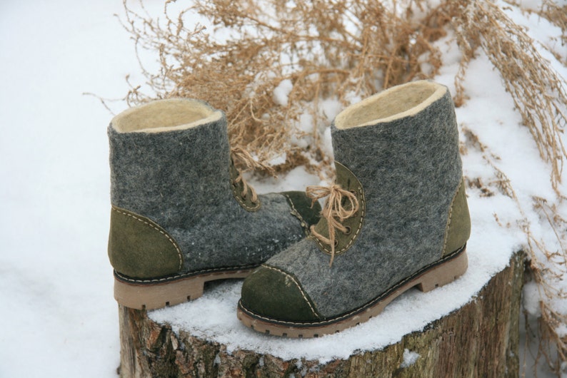 May include: A pair of gray felt boots with green suede accents and brown laces. The boots are sitting on a tree stump covered in snow.