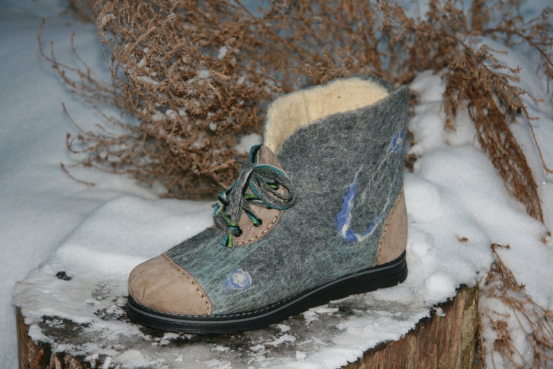 May include: Gray felt boots with a brown leather toe and blue and green laces. The boots are sitting on a snowy surface.
