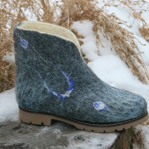 May include: A pair of blue felt boots with a white and blue design. The boots have a brown rubber sole and are sitting on a snowy surface.