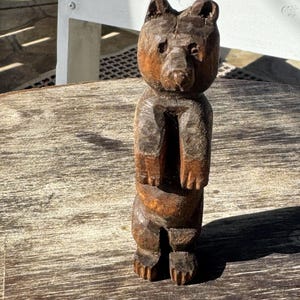 Puede incluir: Figura de oso de madera tallada a mano con un aspecto rústico y texturizado. El oso está representado de pie, con detalles visibles en el pelaje y los rasgos faciales. La madera tiene un tono marrón cálido, lo que sugiere un acabado natural. Un artículo decorativo.