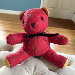 May include: A red quilted teddy bear with a black ribbon around its neck. The bear has a holly and berry pattern and the initials "VB" embroidered on its chest.