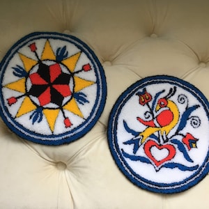 Puede incluir: Dos tapices de pared redondos, blancos, esponjosos, de punto de aguja con bordes azules. El primero presenta un diseño de estallido de estrella rojo, negro y amarillo. El segundo presenta un pájaro amarillo con acentos rojos en una rama con flores rojas y un corazón rojo.