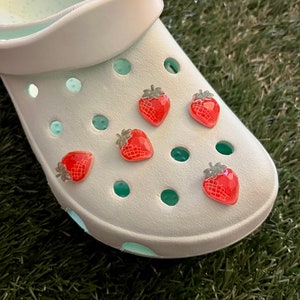 May include: White clogs with five red strawberry charms attached to the holes. The clogs are on a green surface.