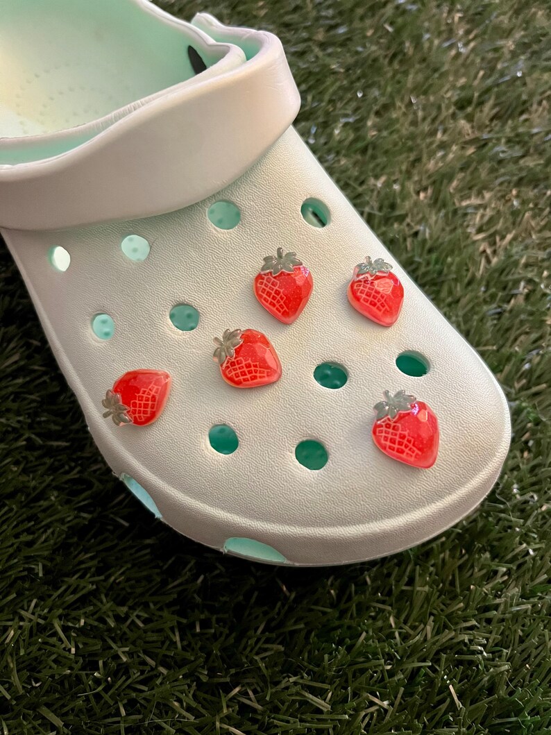 May include: White clogs with five red strawberry charms attached to the holes. The clogs are on a green surface.