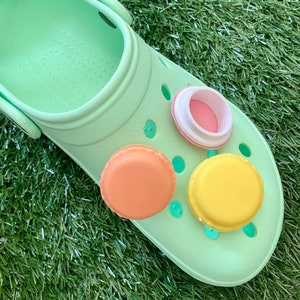May include: Three colorful shoe charms shaped like macarons. The charms are pink, orange, and yellow. The charms are on a green shoe.