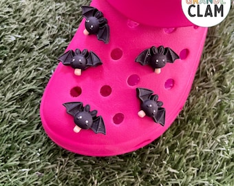 May include: Pink Crocs with black bat popsicle shoe charms. The charms are attached to the shoe with a small hole. The text "THE GRANDE CLAM" is on the side of the shoe.
