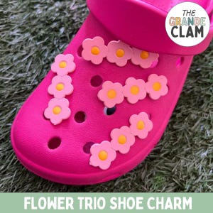May include: Pink shoe with a flower charm design. The charm features three pink flowers with yellow centers. The shoe has the text "THE GRANDE CLAM" on the side.  The shoe is on a green surface.  Flower Trio Shoe Charm.