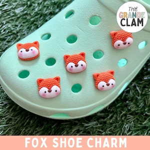 May include: A pair of green Crocs with orange fox shoe charms. The charms are attached to the holes on the shoes. The text "THE GRANDE CLAM" is on the shoe.