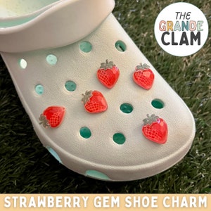 May include: White clog shoe with five red strawberry gem shoe charms. The charms have a silver-colored metal accent. The shoe is on a green grassy surface. The text "THE GRANDE CLAM" is in a circle above the shoe.  "STRAWBERRY GEM SHOE CHARM" is written below the shoe.