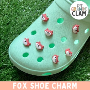 May include: A pair of green clogs with fox shoe charms. The charms are red and have a simple fox design. The clogs have the words "The Grande Clam" on the side.