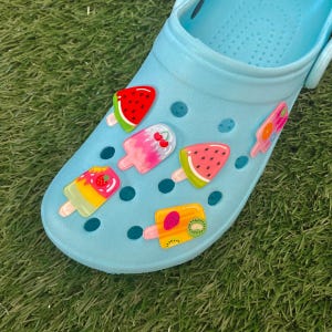 May include: Blue clogs with colourful fruit-shaped charms. The charms are shaped like popsicles and feature watermelon, strawberry, kiwi, and cherry designs.