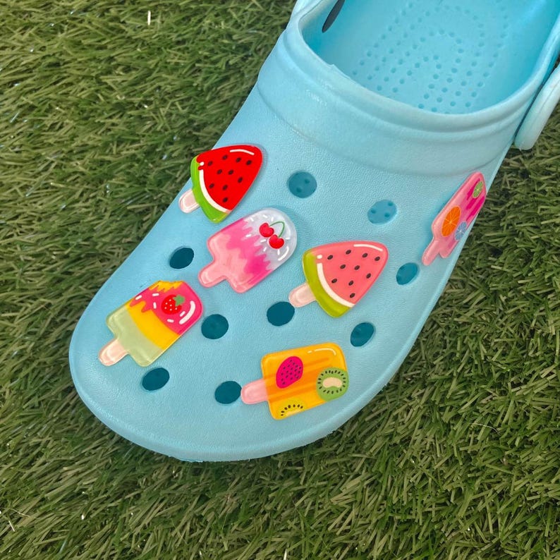 May include: Blue clogs with colourful fruit-shaped charms. The charms are shaped like popsicles and feature watermelon, strawberry, kiwi, and cherry designs.