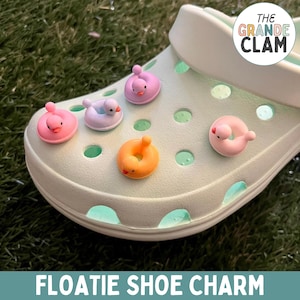 May include: A white pair of clogs with five colorful inflatable pool float shoe charms. The charms are shaped like a duck, a ring, and a flamingo. The charms are attached to the clogs with holes. The text "THE GRANDE CLAM" is on the side of the clogs. The text "FLOATIE SHOE CHARM" is at the bottom of the image.