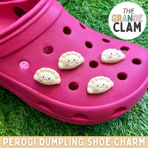 May include: Pink Crocs with four yellow and white perogi dumpling shoe charms. The charms have a smiling face and are attached to the shoe with a small hole. The text "THE GRANDE CLAM" is on a sticker on the shoe. The text "PEROGI DUMPLING SHOE CHARM" is at the bottom of the image.
