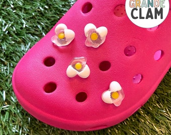 May include: Pink Crocs shoe with four white flower charms with yellow centers. The charms are attached to the shoe. The text "THE GRANDE CLAM" is on a white circle with a rainbow border. The text "BROKEN EGG SHOE CHARM" is at the bottom of the image.