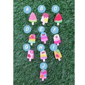 May include: Ten colourful popsicle charms with different fruit designs, labelled A through J. The charms are on a green grassy background.