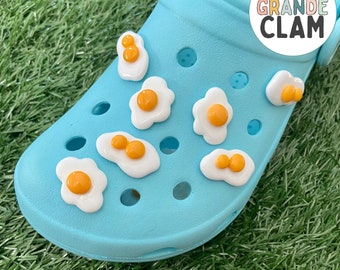 May include: Blue shoe charm with multiple fried egg designs. The shoe charm is made of plastic and has a glossy finish. The text "THE GRANDE CLAM" is printed on the shoe charm.  "FRIED EGG SHOE CHARM" is printed below the shoe charm.