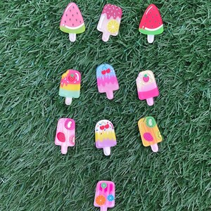 May include: Ten colourful popsicle-shaped charms with various fruit and candy designs. The charms are made of plastic and have a glossy finish.
