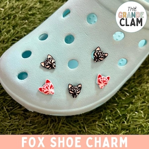 May include: Four fox shoe charms on a light blue shoe. Two charms are black and white, and two charms are red and white. The shoe is on a green grassy surface. The text "THE GRANDE CLAM" is in the upper right corner of the image. The text "FOX SHOE CHARM" is at the bottom of the image.