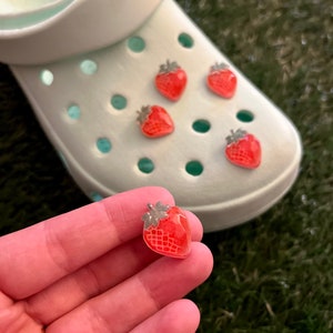 May include: Five red strawberry shoe charms with green leaves. The charms are on a white shoe with holes. One charm is being held in a hand.