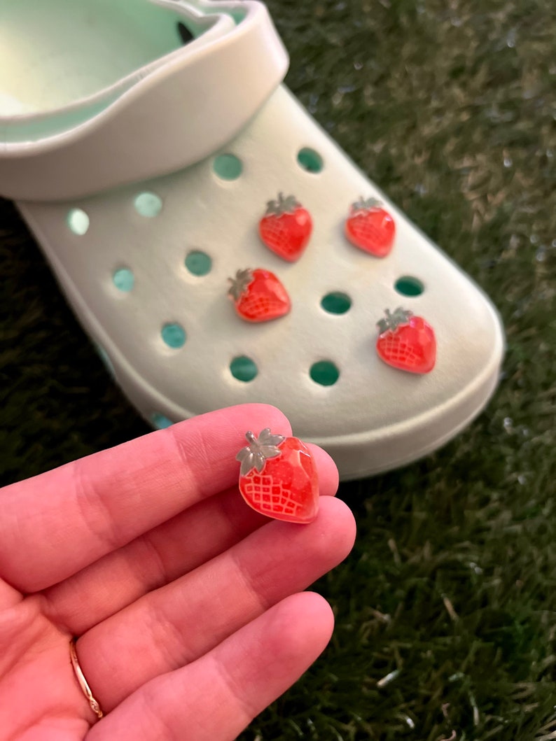 May include: Five red strawberry shoe charms with green leaves. The charms are on a white shoe with holes. One charm is being held in a hand.