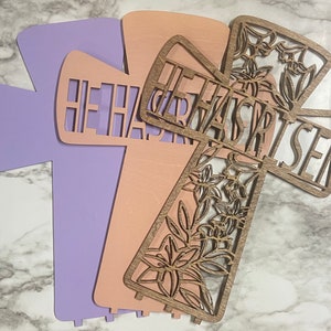 May include: Three wooden crosses with the words "He Has Risen" cut out. The crosses are layered on top of each other, with the top cross being a floral design. The crosses are painted in shades of pink and purple.