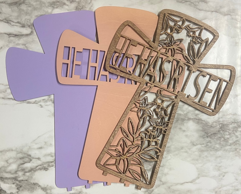 May include: Three wooden crosses with the words "He Has Risen" cut out. The crosses are layered on top of each other, with the top cross being a floral design. The crosses are painted in shades of pink and purple.