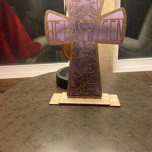 May include: A wooden cross with a purple floral design and the words "He Has Risen" on the front. The cross is standing on a wooden base.