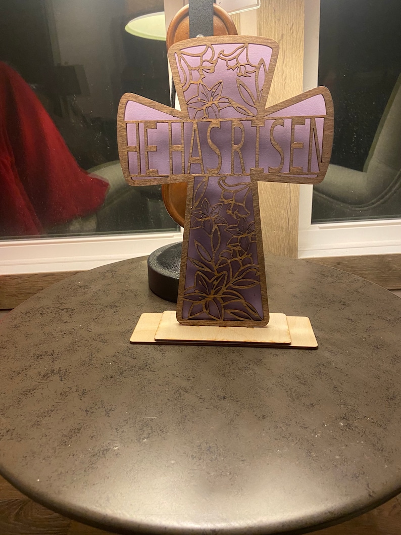 May include: A wooden cross with a purple floral design and the words "He Has Risen" on the front. The cross is standing on a wooden base.