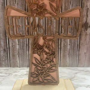 May include: A wooden cross with a floral design and the words "He Has Risen" cut out. The cross is painted a light pink color and has a wooden base.