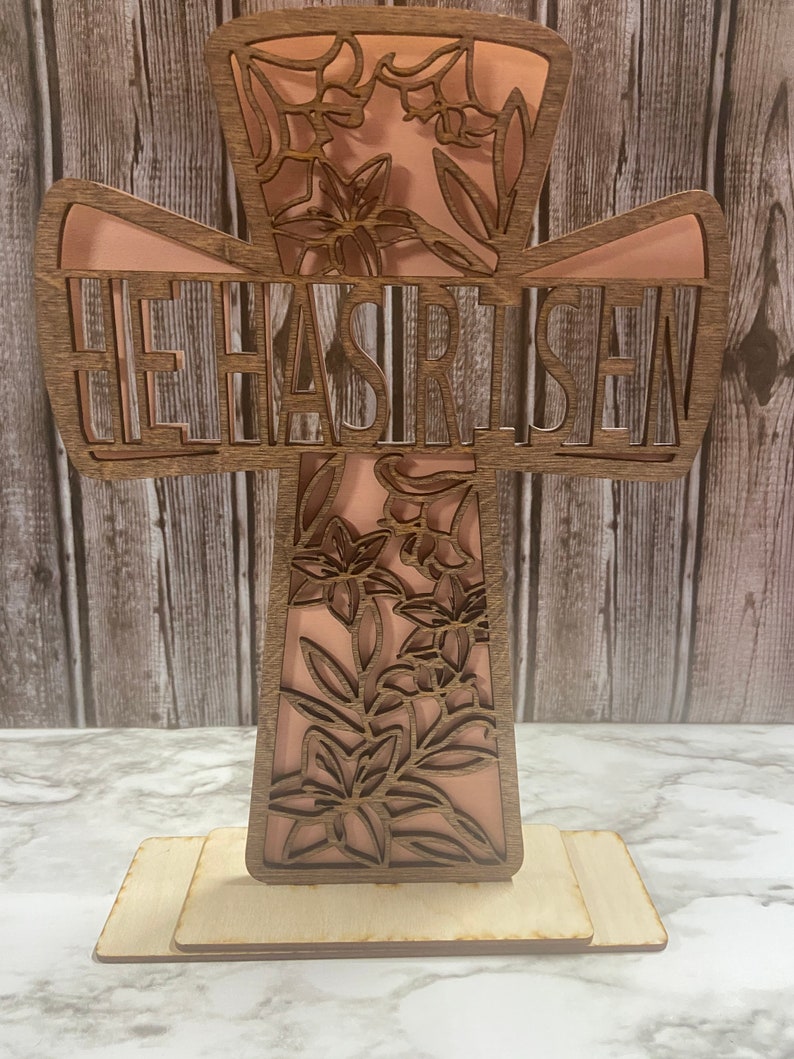 May include: A wooden cross with a floral design and the words "He Has Risen" cut out. The cross is painted a light pink color and has a wooden base.