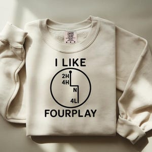 May include: Cream-coloured sweatshirt with the text "I LIKE FOURPLAY" in black. The design includes a gear shift graphic. The sweatshirt has a crew neck and long sleeves with ribbed cuffs.