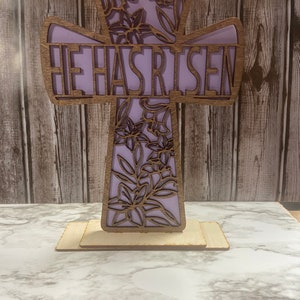 May include: A wooden cross with a purple background and floral design. The words "He Has Risen" are cut out of the cross.