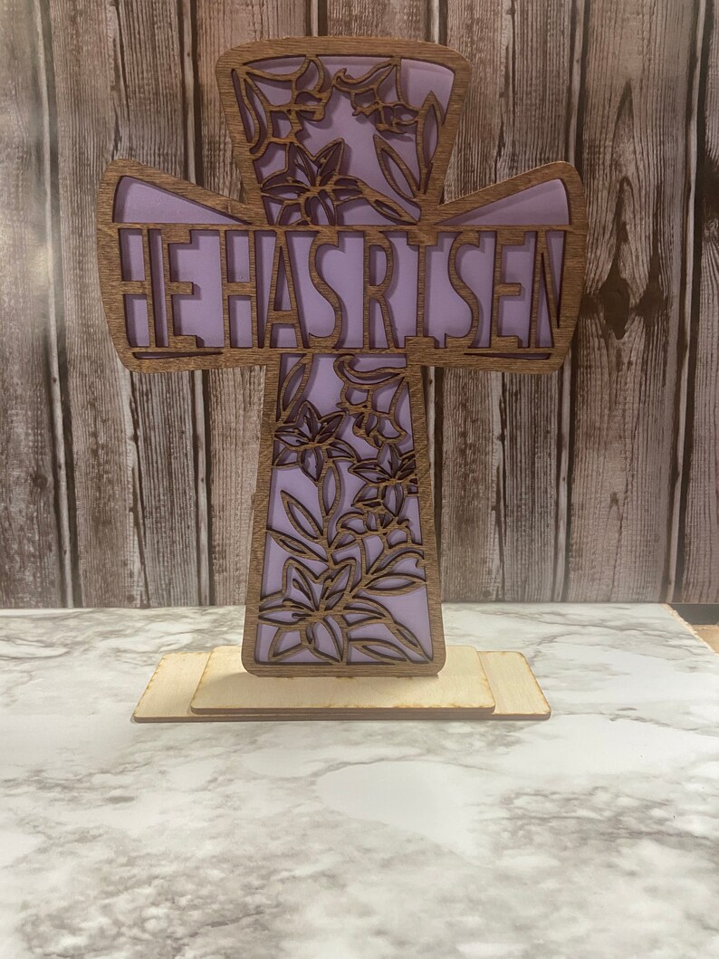 May include: A wooden cross with a purple background and floral design. The words "He Has Risen" are cut out of the cross.