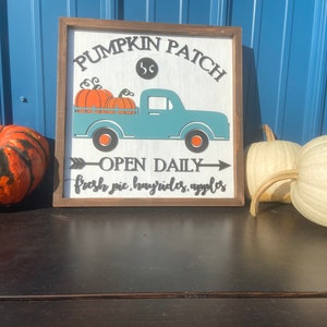 May include: A wooden sign with a light blue truck carrying orange pumpkins, reading "Pumpkin Patch, Open Daily". A large orange and black pumpkin and two white pumpkins are next to the sign, on a dark surface.