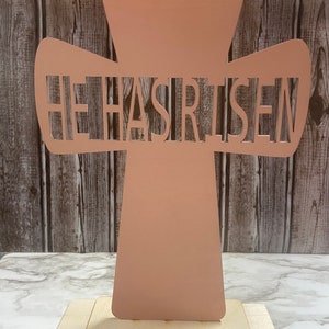 May include: A light pink wooden cross with the words "He Has Risen" cut out. The cross is standing on a wooden base.
