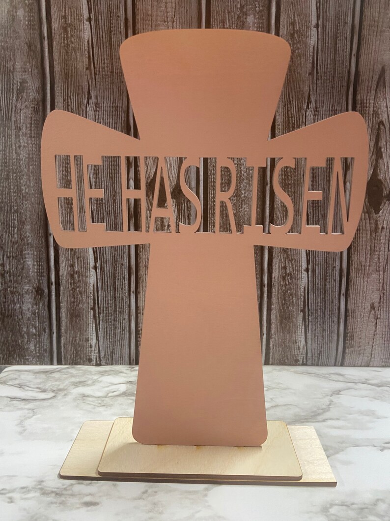 May include: A light pink wooden cross with the words "He Has Risen" cut out. The cross is standing on a wooden base.