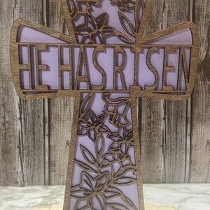 May include: A wooden cross with a floral design and the words "He Has Risen" in a purple font. The cross is standing on a wooden base.