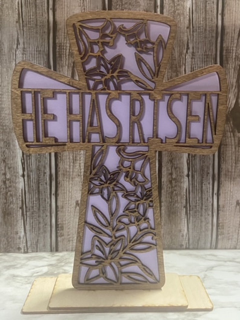 May include: A wooden cross with a floral design and the words "He Has Risen" in a purple font. The cross is standing on a wooden base.