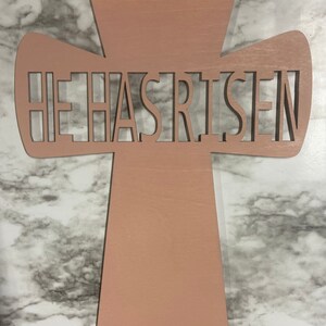May include: A wooden cross with the words "He Has Risen" cut out in the center. The cross is painted a light pink color.