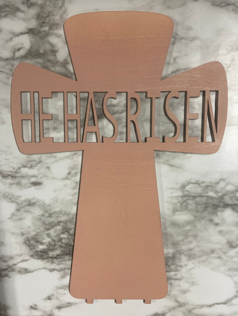 May include: A wooden cross with the words "He Has Risen" cut out in the center. The cross is painted a light pink color.