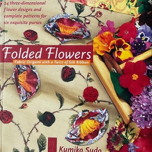 May include: A book titled "Folded Flowers" with fabric origami designs. The cover features fabric flower purses and the text "Fabric Origami with a Twist of Silk Ribbon." The image includes real flowers in a wooden tray.