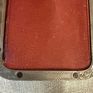 May include: Close-up of a rectangular, reddish-brown leather item with a silver-colored metal frame. The frame has decorative scrollwork and small screws. The leather appears worn, suggesting age or use. The item is set against a textured, off-white background.