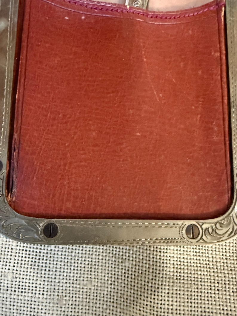 May include: Close-up of a rectangular, reddish-brown leather item with a silver-colored metal frame. The frame has decorative scrollwork and small screws. The leather appears worn, suggesting age or use. The item is set against a textured, off-white background.