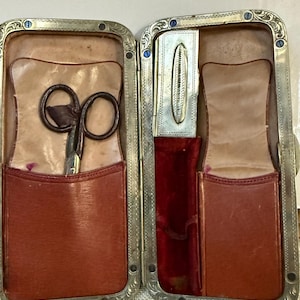 May include: Open antique travel grooming kit with a silver-toned frame and decorative edges. The interior features brown leather pockets, a pair of scissors, and a small silver case. The lining is tan leather and red velvet.